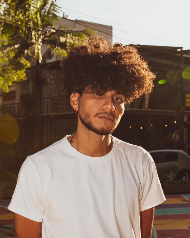 Man with curly hair in a plain white t-shirt on a sunny street, with palm fronds and buildings behind
