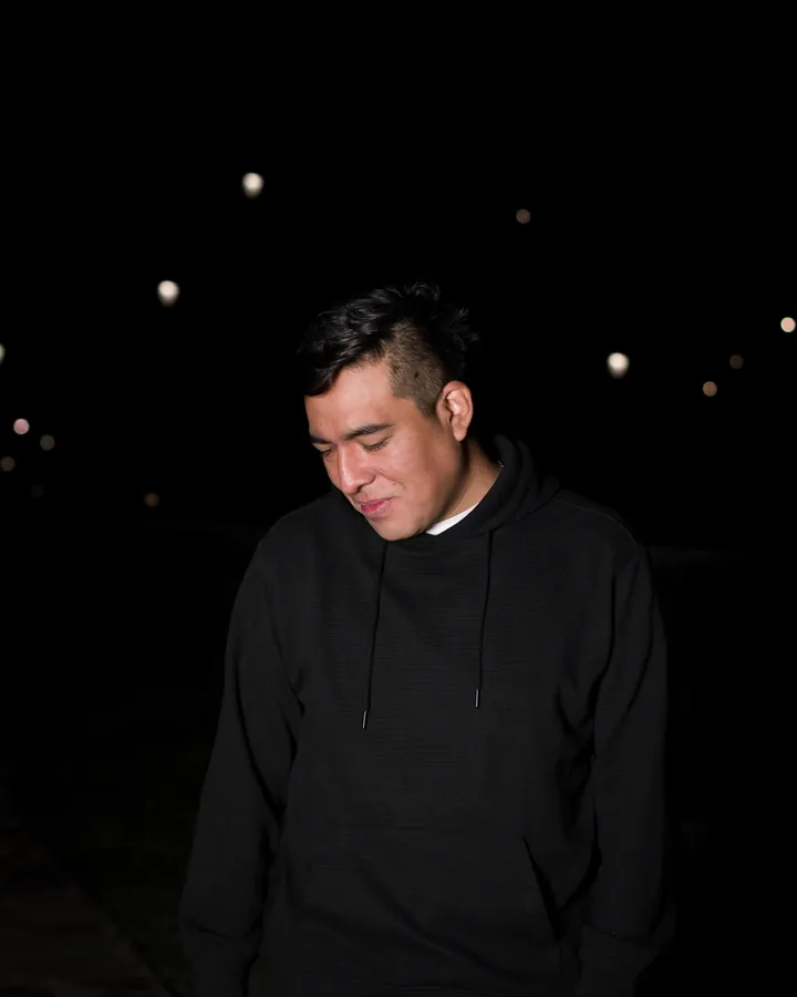 Man in a plain black hoodie looking down, outdoors at night with blurred lights behind