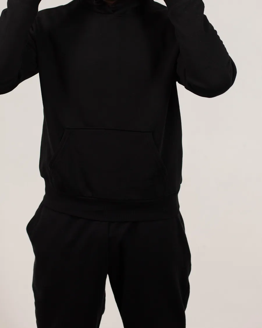 Plain black pullover hoodie with a kangaroo pocket, worn with black sweatpants, cropped below the face against a light studio wall