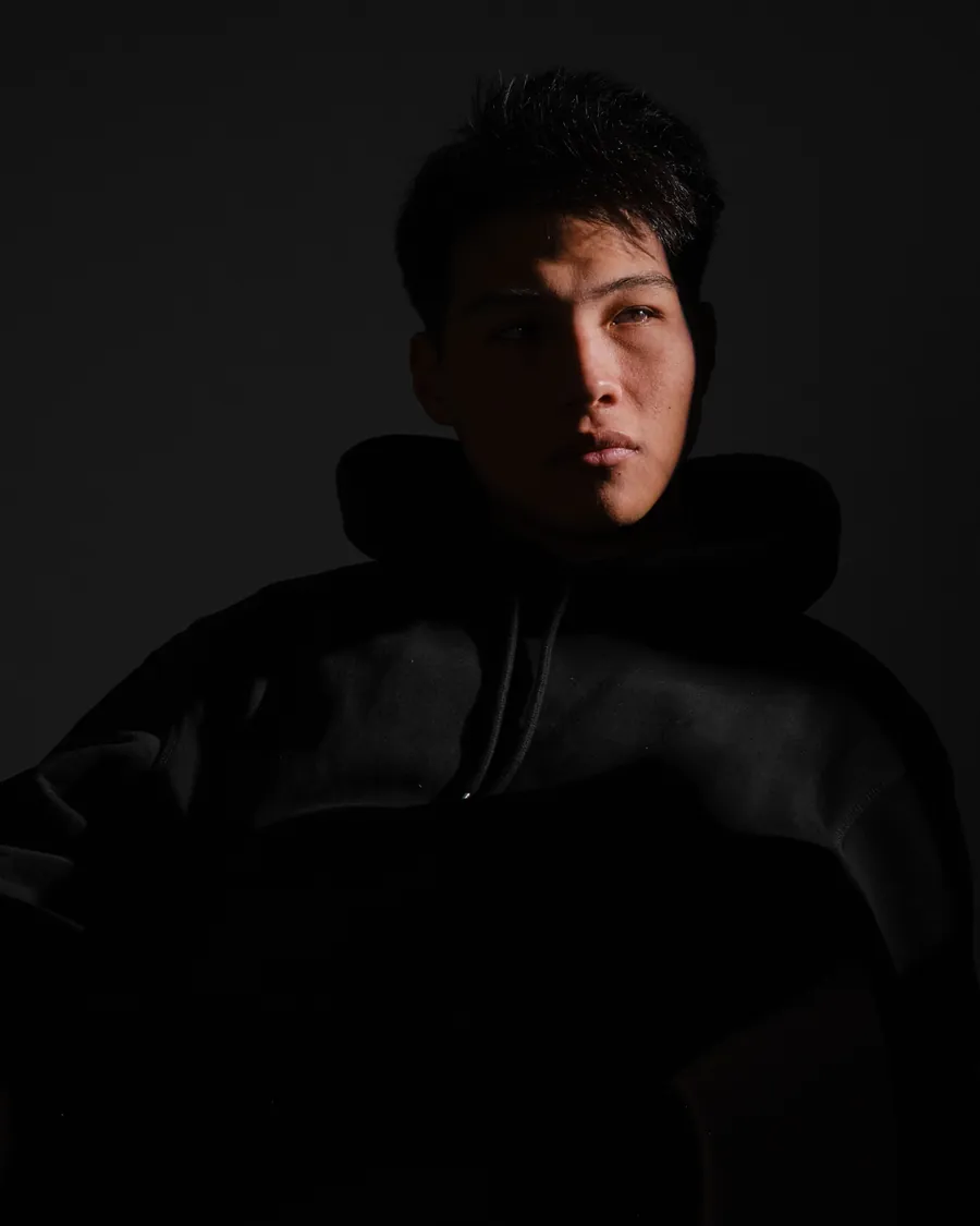 Young man in an oversized black hoodie, side-lit against a black background