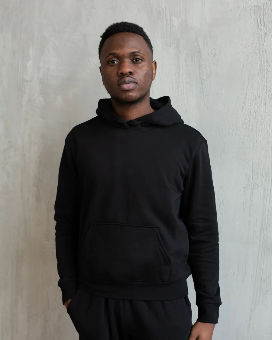 Man in a plain black hoodie and black sweatpants standing against a grey concrete wall