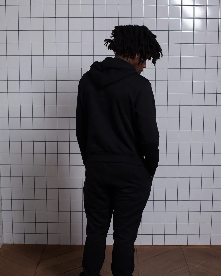 Back view of a person with locs in a black hoodie and black joggers in a white-tiled room
