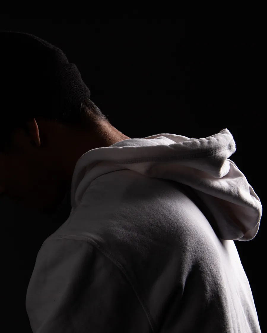 Person in a white hoodie and black beanie with head bowed, lit against a black background
