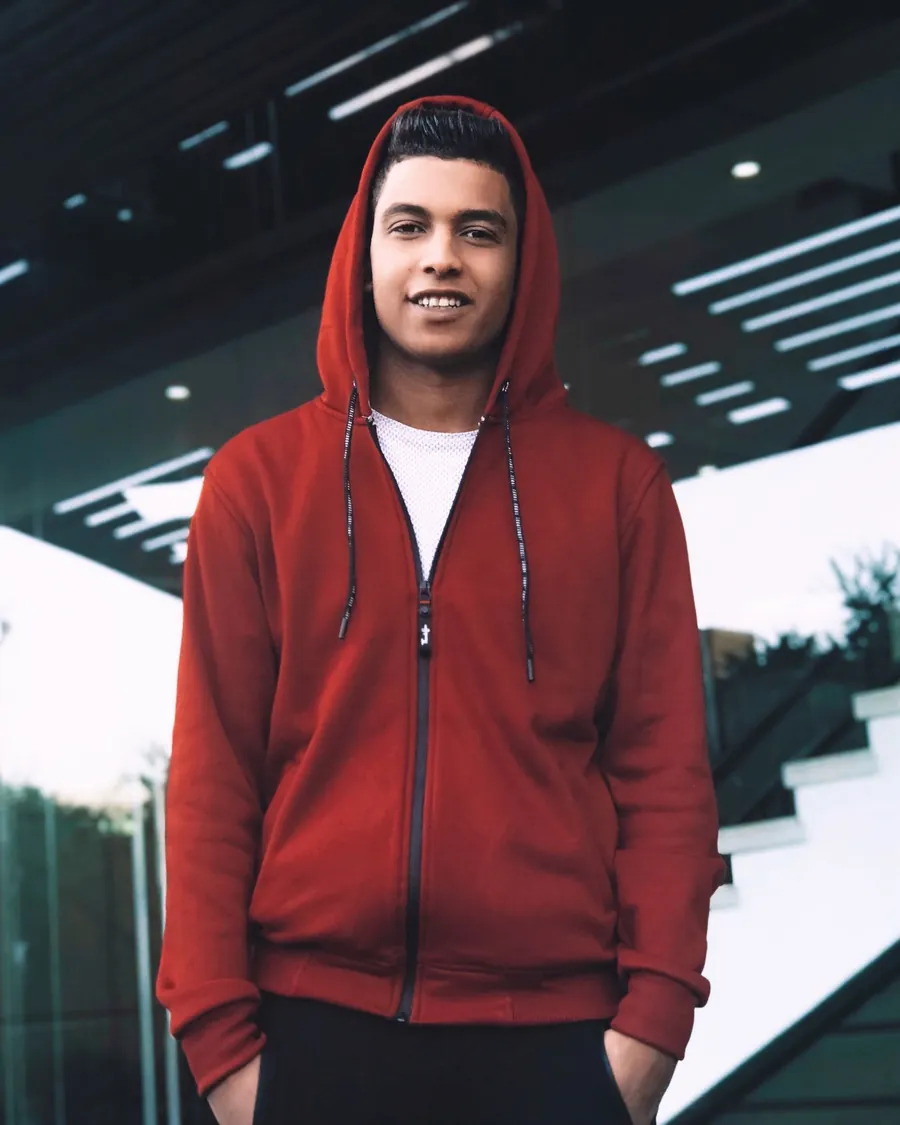 Smiling young man in a red zip-up hoodie over a white tee, outside a glass building