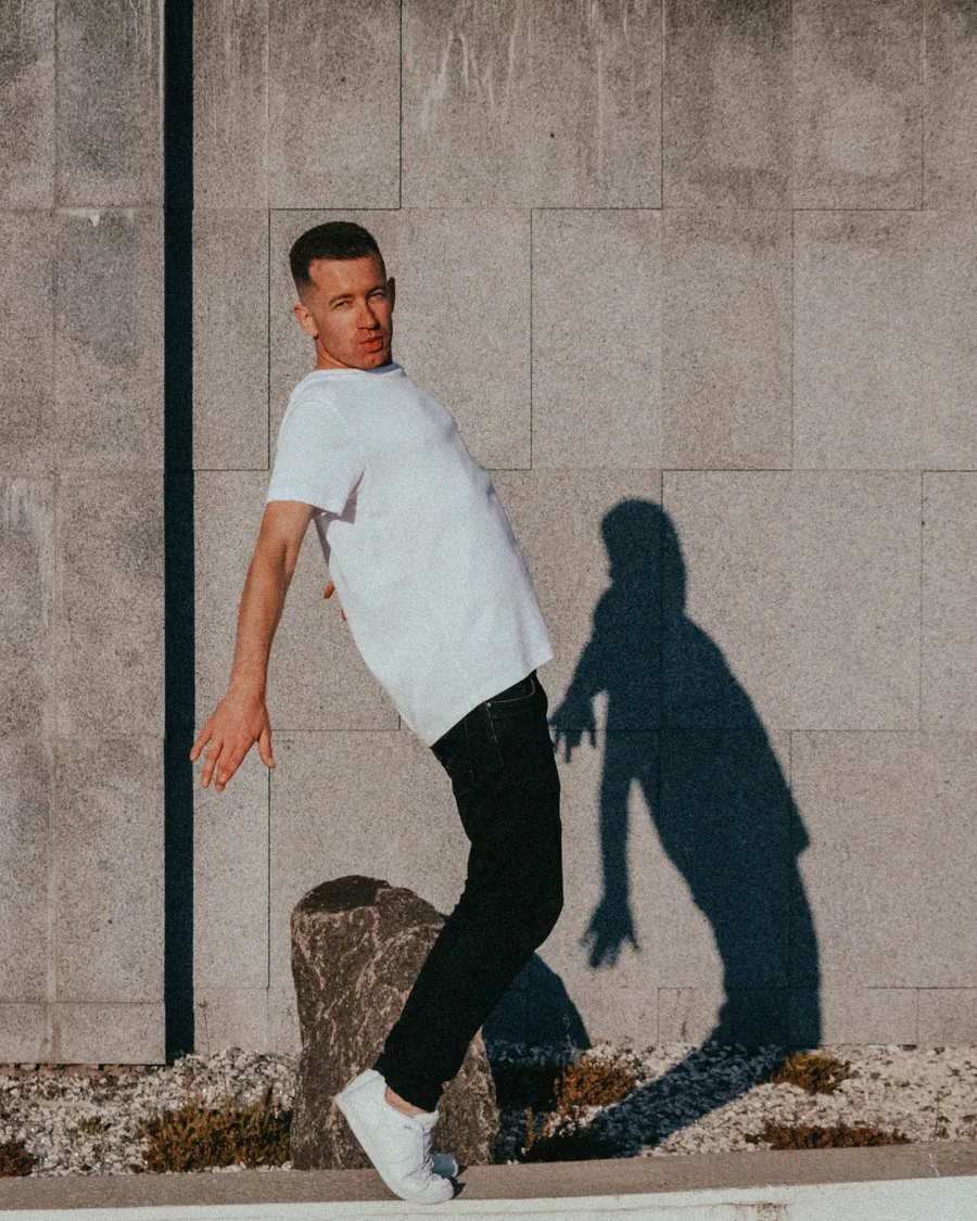 Man in a plain white t-shirt, black jeans and white sneakers leaning back against a sunlit stone wall, his shadow beside him