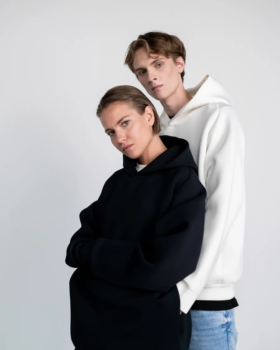 Woman in a black hoodie and man in a white hoodie posing together against a light grey studio wall