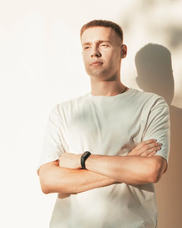 Man in a plain cream-white t-shirt with arms crossed against a sunlit wall with leaf shadows