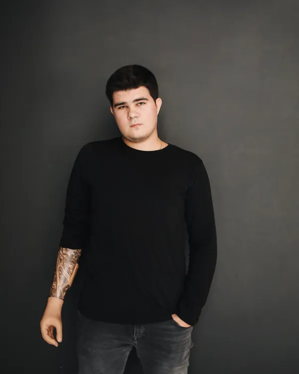 Man in a plain black long-sleeve t-shirt and grey jeans against a dark grey wall