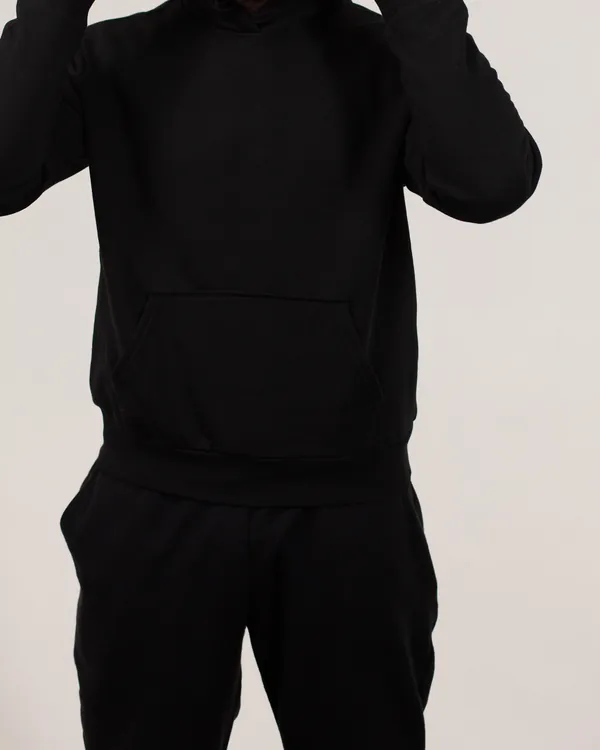 Plain black pullover hoodie with a kangaroo pocket, worn with black sweatpants, cropped below the face against a light studio wall