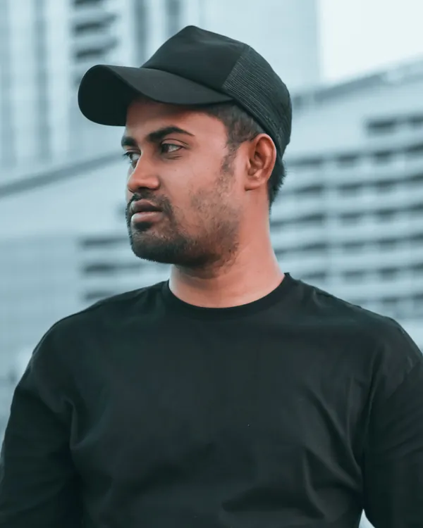 Man in a plain black cap and black t-shirt in front of blurred city buildings