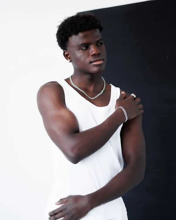 Young man in a white tank top and silver chain against a white and black studio backdrop