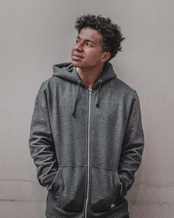 Young man in a charcoal grey zip-up hoodie with his hands in the pockets, against a pale concrete wall