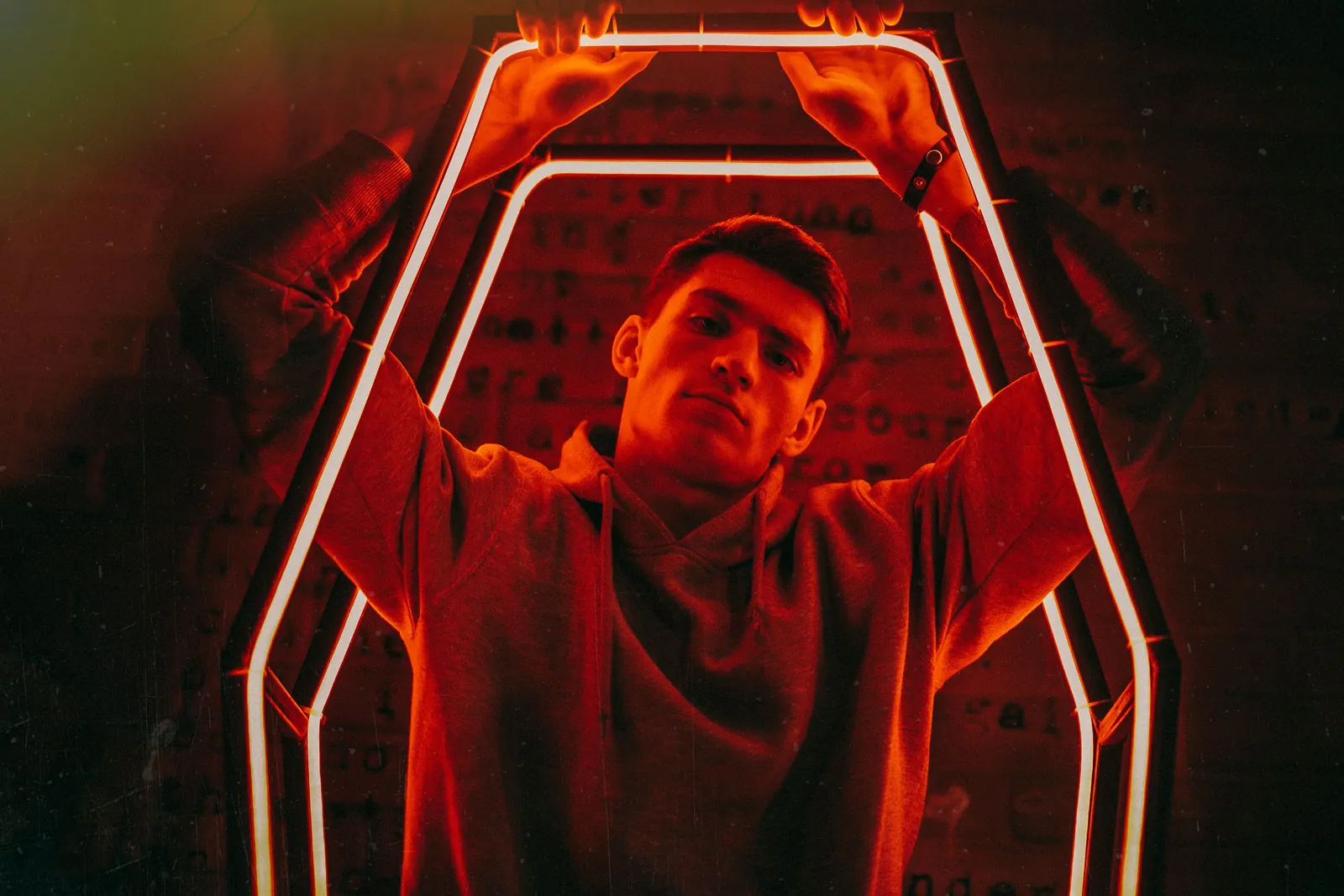 Young man in a hoodie holding up a hexagonal red neon light frame, the whole scene lit red