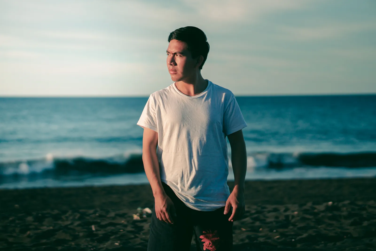 Young man in a plain white t-shirt and dark shorts standing on a beach in low golden sun, with ocean waves behind